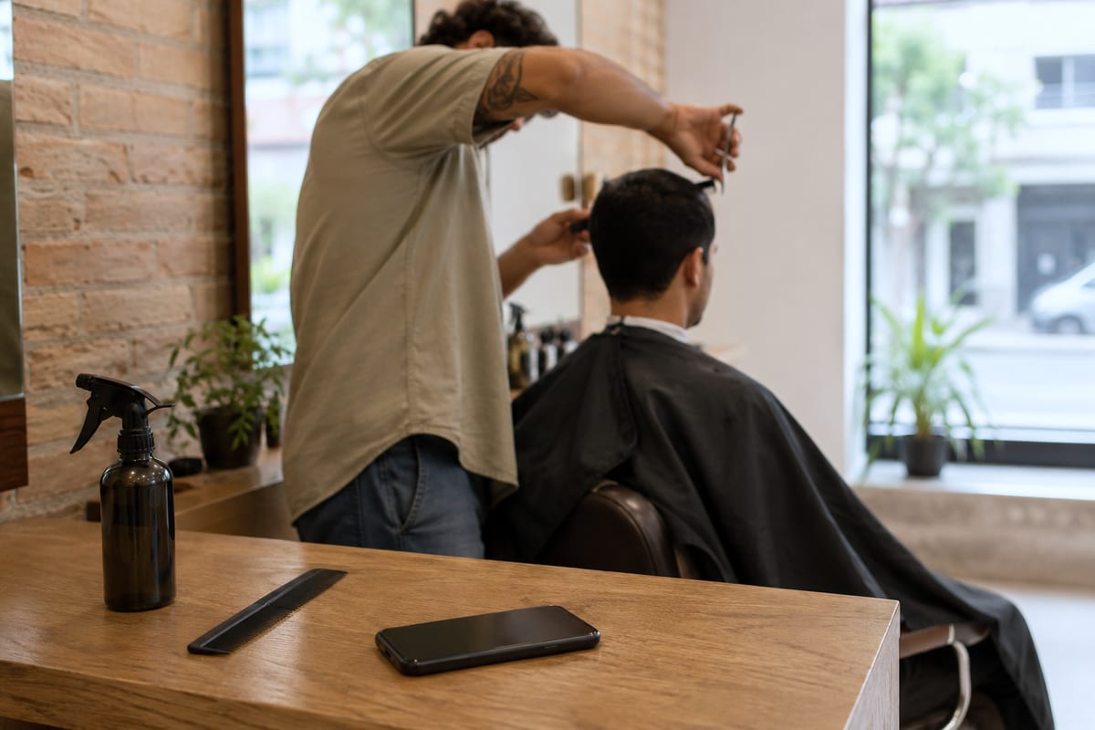 Barbeiro de costas cortando o cabelo de um cliente, com o celular apoiado na bancada